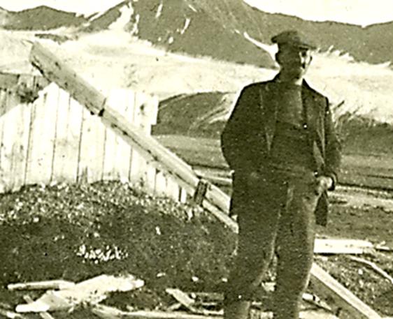 Mann med caps og ullklær står foran en enkel og lav trehytte med fjell i bakgrunnen.