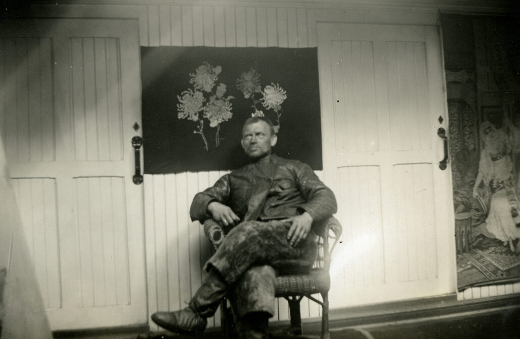 Mann i skitne arbeidsklær sitter på en stol med bena i kors. det er to dører på veggen bak han og et bilde av blomster