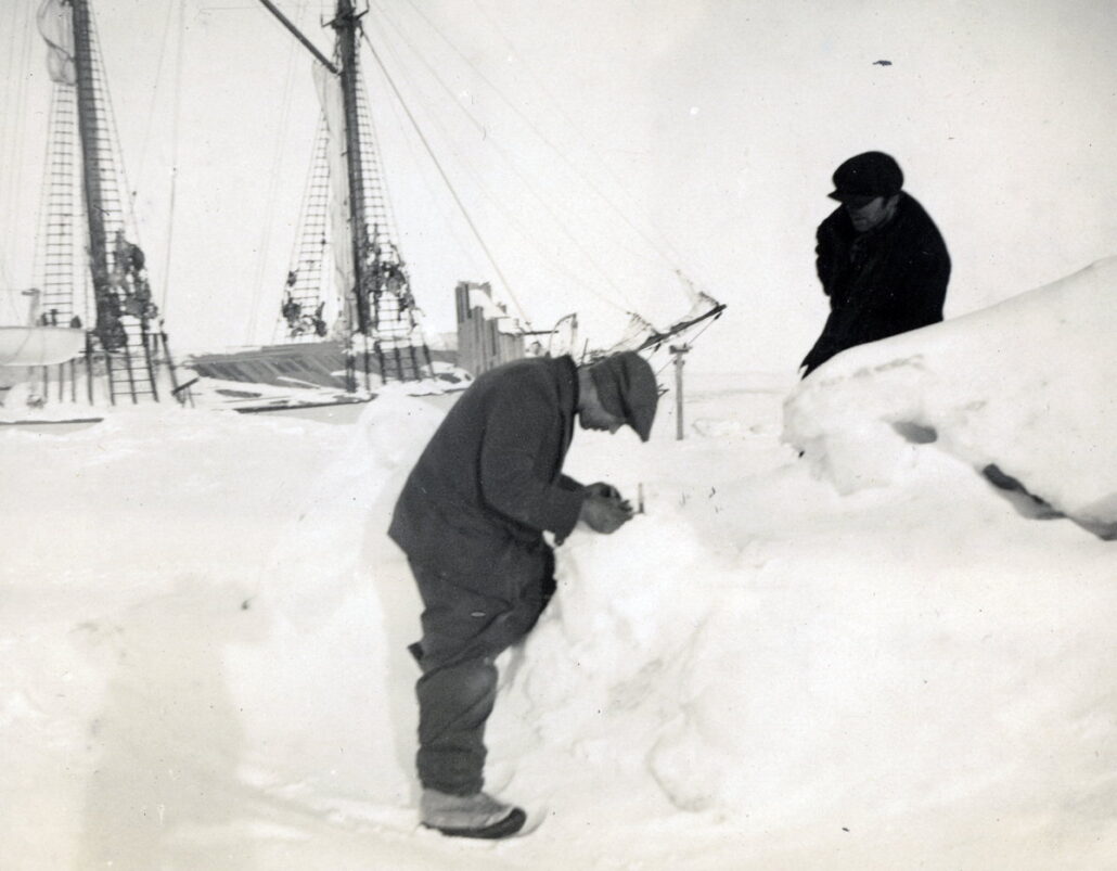 To menn arbeider med utstyr i snø. En stor båt ligger i bakgrunnen