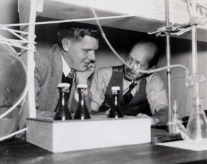 To menn hviler armene på et slags bord med hyller, flasker, ledninger og utstyr rundt
