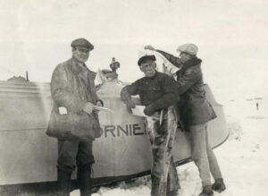 Tre menn står ved en liten flybåt. De har six pence luer.