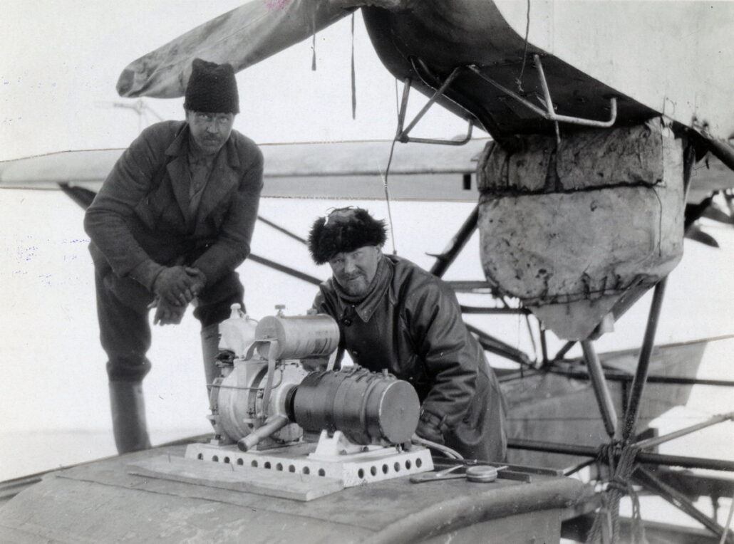 To menn arbeidwet på en maskindel under et lite propellfly, de ser mot fotografen