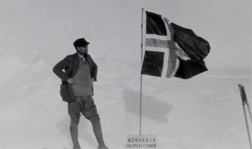 Mann i ullnikkers, lue og åpen jakke står og ser på et vaiende norsk flagg