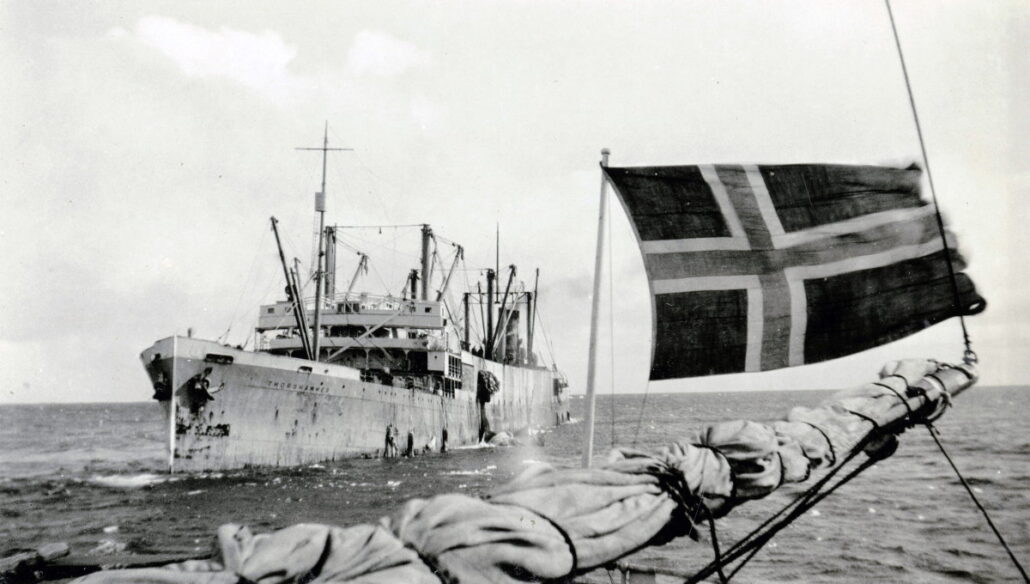 Stor båt på sjøen i forgrunnen masten av en annen båt og det norske flagg