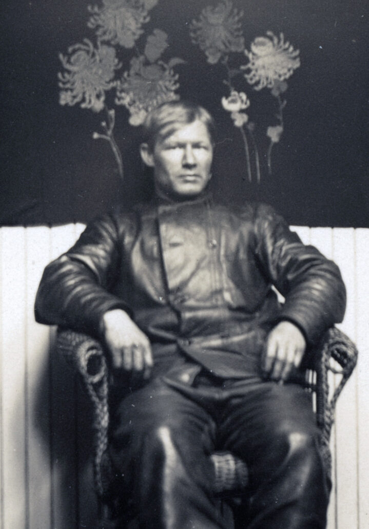 Mann i sort lærjakke og -bukse sitter i en rottingstol, bak han henger et svart veggteppe med blomster