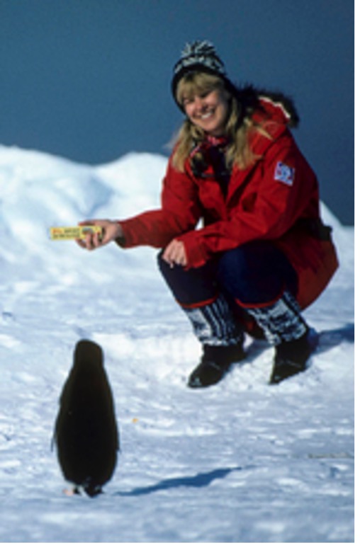 Smilende kvinne sitter på huk foran en pingvin og holder en sjokolade i handa. Hun har en røs jakke med norsk flagg på armen