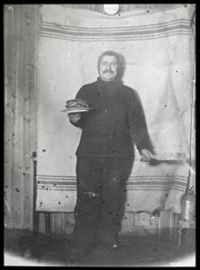 Mann med bart og lue holder en stabel pannekaker i høyre hånd og stekespade i venstre hånd