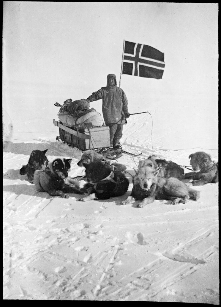 Mann i anorakk står ved slede med norsk flagg. Han har hundepisk i hånden og foran han ligger ni hunder i sneen