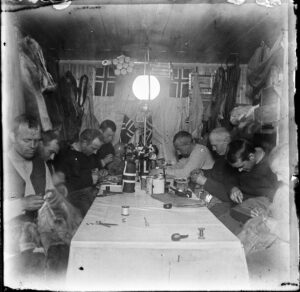 Åtte menn sitter og arbeider konsentrert med hver sitt handarbeide ved et bord i etrangt rom med masse skinnklær og to norske flagg hengende på veggen