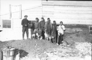 Gruppebilde av seks menn som står oppstilt utenfor en husvegg. Fem hunder står og sitter foran dem.