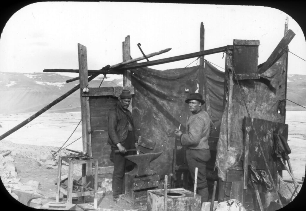 To menn i ullklær står i et primivt oppsatt verksted utendørs med presenning og noen planker som vegger