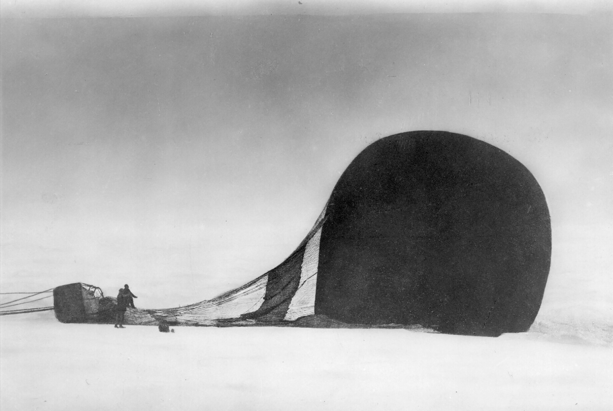 Sort-hvitt bilde med en stor luftballong liggende på sne, to menn står ved siden