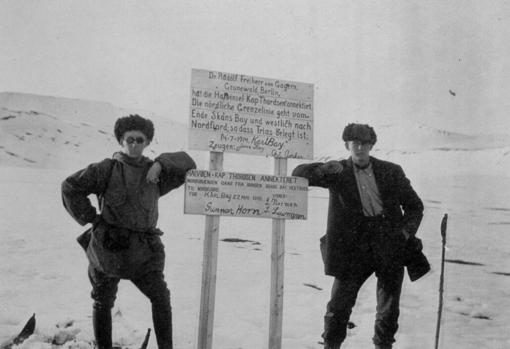 To menn står og hviler albuen mot et skilt med to tavler som står plassert i snøen med to stolper