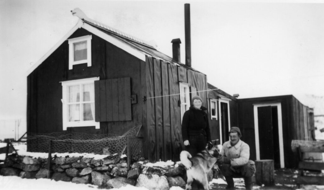 sort-hvit foto med hytta, ei kvinne som står og en mann som sitter ved siden av den. Mannen leker koser med hund.