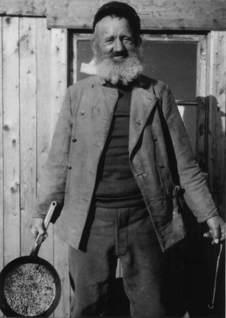 Mann med skjegg, caps på hodet og ukneppet jakke : Gustav Lindquist står foran bygning, holder i stekepanne.
