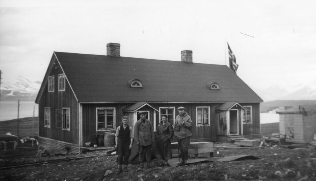 tre menn og en kvinne posrer foran trebygning med to piper og et norsk flagg vaiende