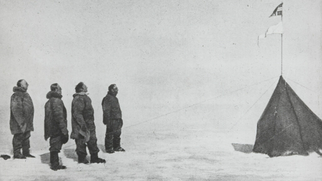 Fire menn med lua i handa står ved en telt med norsk flagg og et hvitt flagg på snedekket bakke