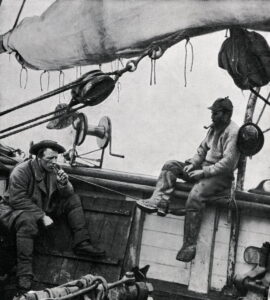 To menn sitter på et båtsdekk og røyker pipe. De ahar på seg ullklær og hatt/caps