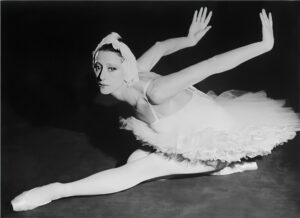Ballettdanserinne sitter på scene med armen bak og opp og venstre fot foran seg.Hun har hvit kjole, strømpebukse og hodepynt