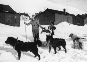 Foran i bildet hunder fastspent på rekke og rad, hundeslede. Bak hundene kvinne på ski, 2 barn, den ene sitter på spark eller slede, og den andre står bak sleden. I bakgrunnen bebyggelse med hus og bygninger. Snø.