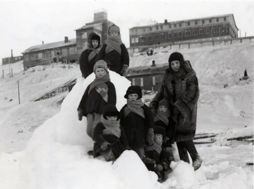 Gruvearbeiderbarn kledd i varme vinterklær leker i snøen. I bakgrunnen ses bebyggelse med hus og bygninger.