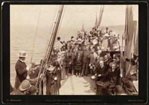 Stor gruppe med pent kledde menn og kvinner står samlet på en båt. Mange menn hilser med hattene