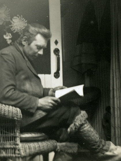 Mann sitter på stol og leser en bok. Foran han står en ovn og bak han er en åpen skyvedør. På veggen bak henger et tøystykke med store blomster