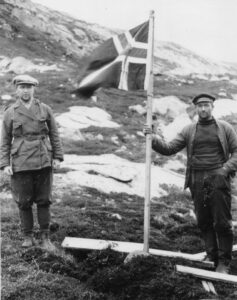 To menn i ullklær og six pence luer. den ene holder i et norsk flagg som står på bakken.