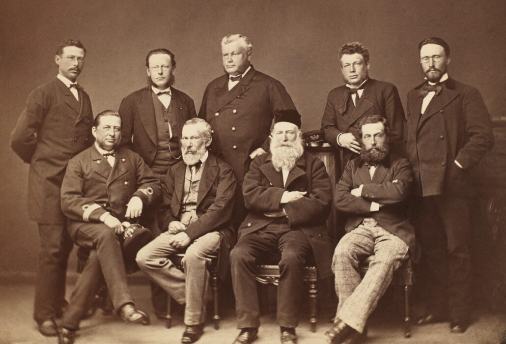 Gruppebilde av ni menn i dress, de fire fremste sitter